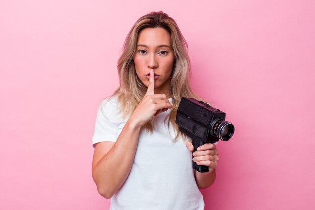 Photo young australian woman filming with a vintage video camera isolated keeping a secret or asking for silence.