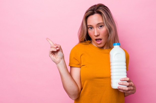 側面を指しているピンクの背景に分離されたミルクのボトルを保持している若いオーストラリア人女性