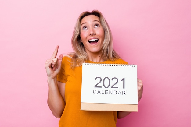 口を開けて逆さまを指しているピンクの背景で隔離のカレンダーを保持している若いオーストラリア人女性。
