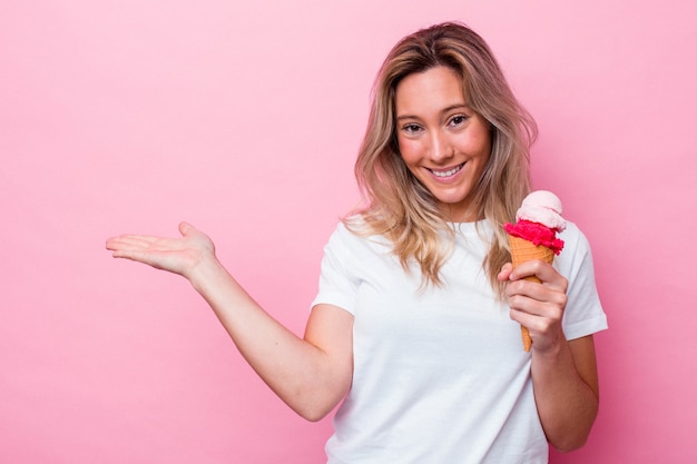 ピンクの背景に分離されたアイスクリームを保持し、手のひらにコピースペースを示し、腰に別の手を保持している若いオーストラリア人女性。