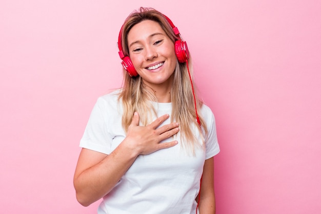 Young australian woman listening music isolated on pink background laughs out loudly keeping hand on chest.