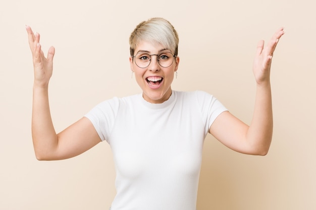 Young authentic natural woman wearing a white shirt receiving a pleasant surprise, excited and raising hands.