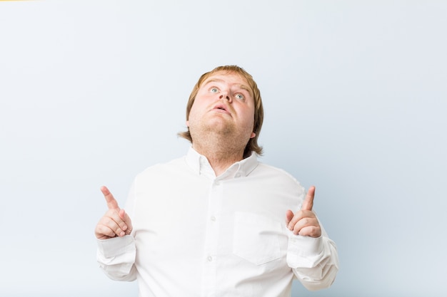 Young authentic redhead fat man pointing upside with opened mouth.