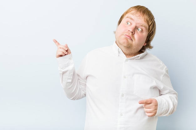 Young authentic redhead fat man shocked pointing with index fingers to a copy space.