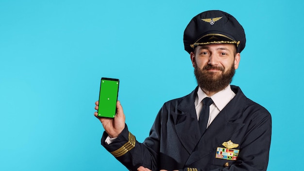 Giovane aviatore che usa lo schermo verde vestito da capitano, mostrando smartphone con display mockup isolato in studio. pilota di volo professionale che tiene il telefono con modello chroma key vuoto.