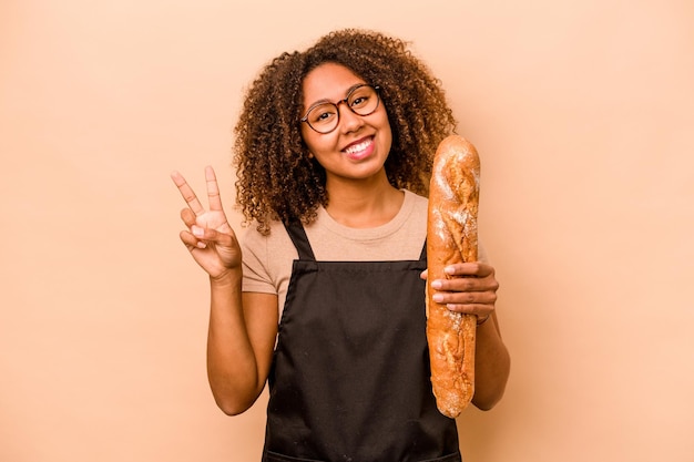 Young baker African woman holding a loaf of bread isolated on beige background joyful and carefree showing a peace symbol with fingers