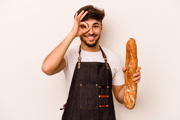 Молодой пекарь, латиноамериканец, изолированный на белом фоне, взволнован, держа хорошо жест на глазу
