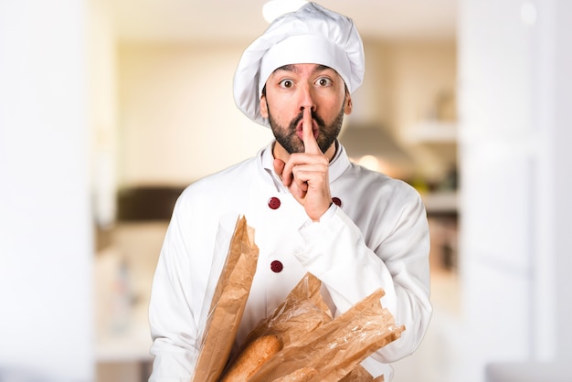 Young baker holding some bread and making silence gesture in the kitchen
