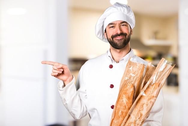 Young baker holding some bread and pointing to the lateral in the kitchen