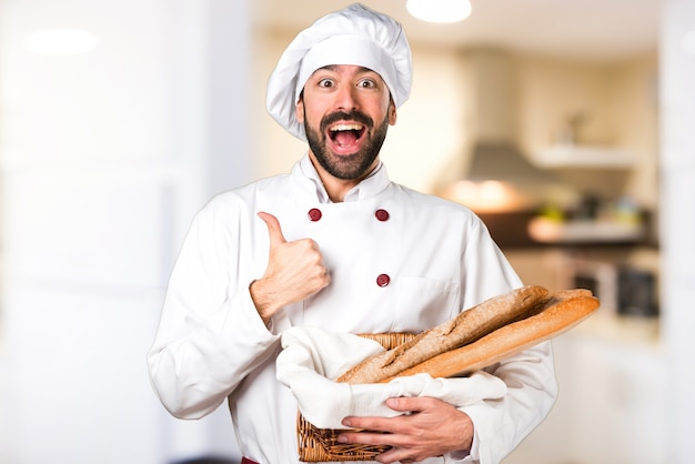 Young baker holding some bread and with thumb up in the kitchen