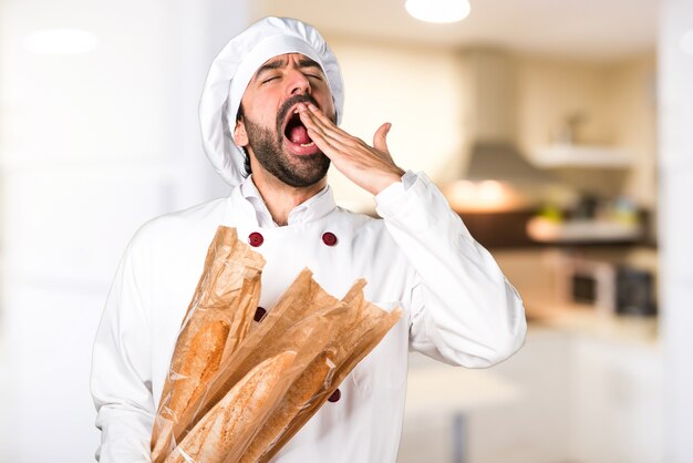 Young baker holding some bread and yawning in the kitchen