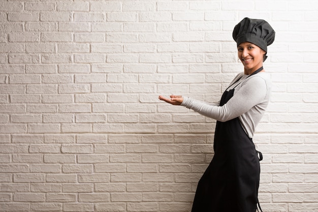 Young baker indian woman against a bricks wall holding something with hands