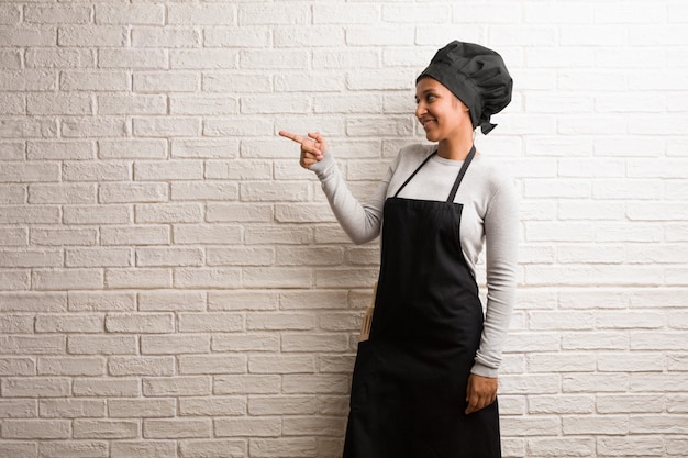 Young baker indian woman against a bricks wall pointing to the side.