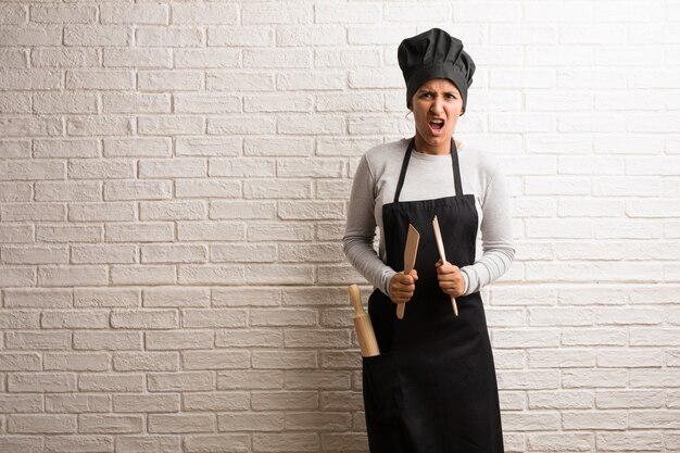 Young baker indian woman against a bricks wall very angry and upset, very tense.