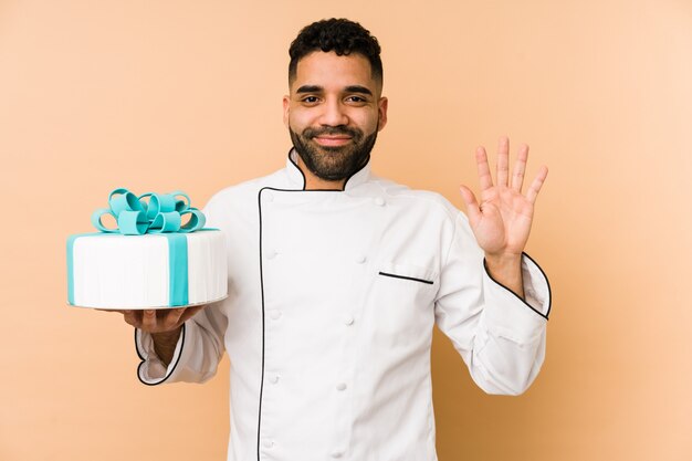 Young baker man holding a cake isolated smiling cheerful showing number five with fingers