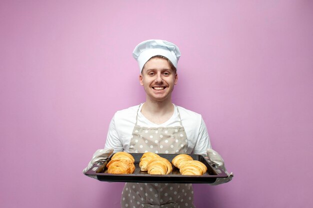 Foto giovane fornaio in uniforme che tiene croissant appena sfornati e sorride su sfondo rosa