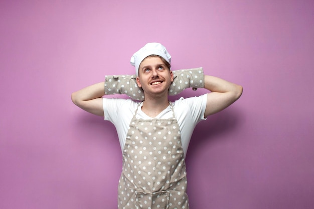 Young baker in uniform put his hands behind his head and rests on pink background