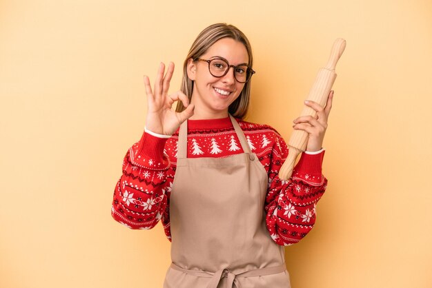 黄色の背景に分離されたクリスマスのクッキーをやっている若いパン屋の女性は陽気で自信を持ってOKジェスチャーを示しています。