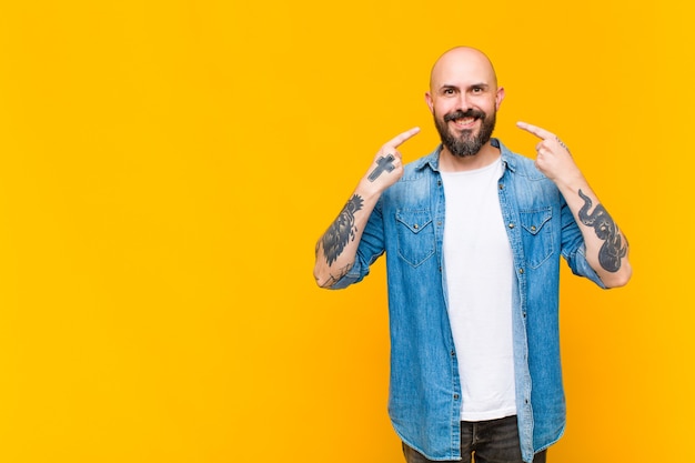 Young bald and bearded man smiling confidently pointing to own broad smile, positive, relaxed, satisfied attitude