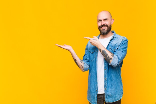 Young bald and bearded man smiling, feeling happy, carefree and satisfied, pointing to concept or idea on copy space on the side