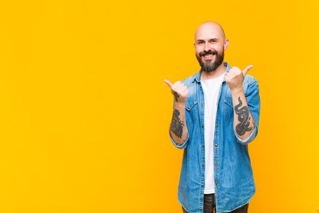 Young bald and bearded man smiling joyfully and looking happy, feeling carefree and positive with both thumbs up