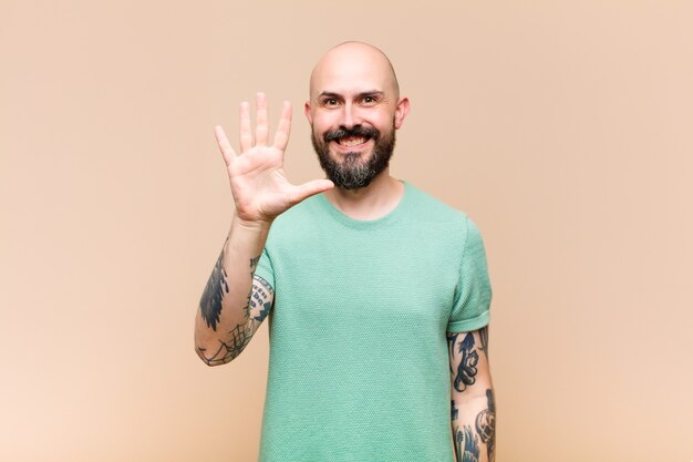 Young bald and bearded man smiling and looking friendly, showing number five or fifth with hand forward, counting down