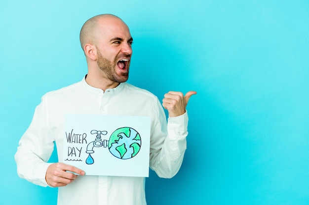 Young bald man celebrating world water day isolated on blue wall points with thumb finger away, laughing and carefree