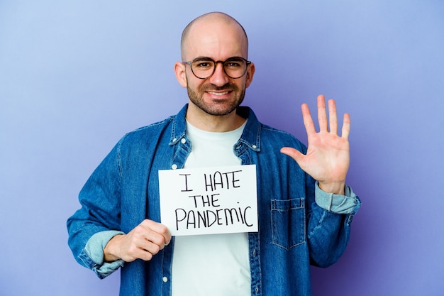 Young bald man holding a I hate the pandemic placard isolated on blue wall smiling cheerful showing number five with fingers