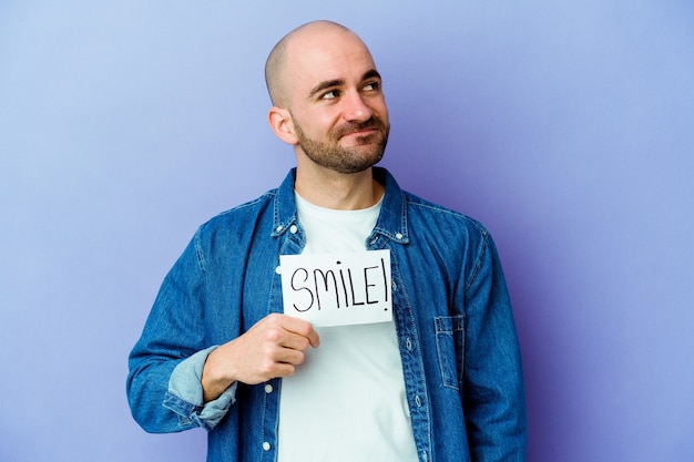 Young bald man holding a smile placard isolated wall dreaming of achieving goals and purposes