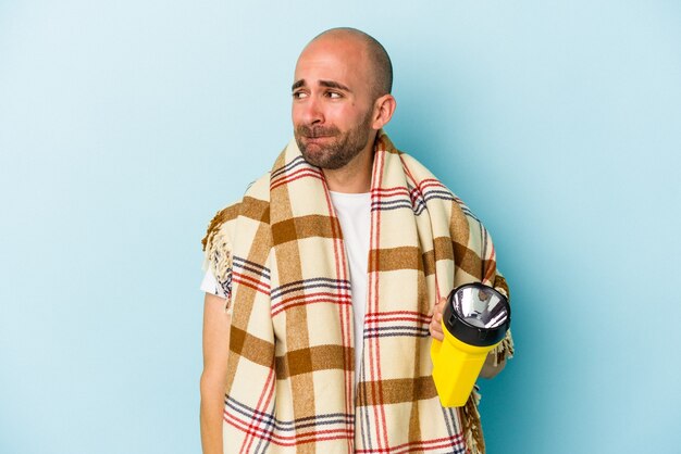 Young bald man holding vintage lantern isolated on blue background  confused, feels doubtful and unsure.