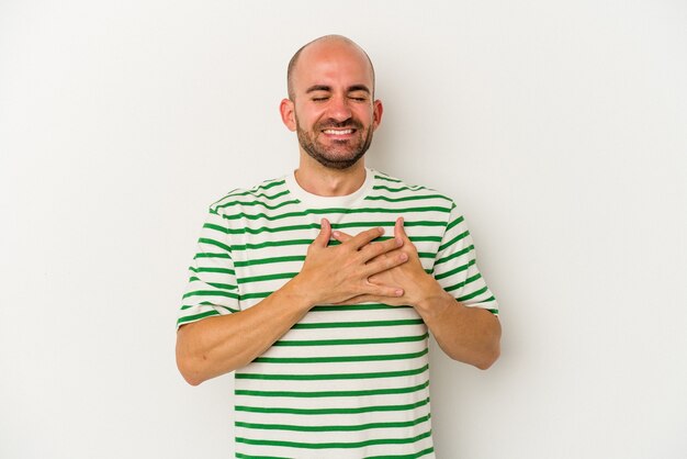 Young bald man isolated on white background laughing keeping hands on heart, concept of happiness.