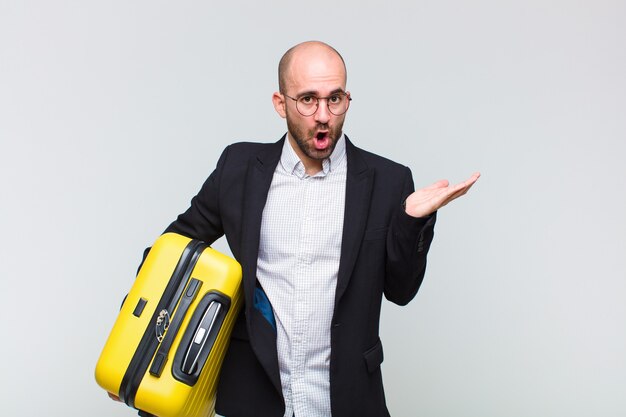 Young bald man looking surprised and shocked, with jaw dropped holding an object with an open hand on the side