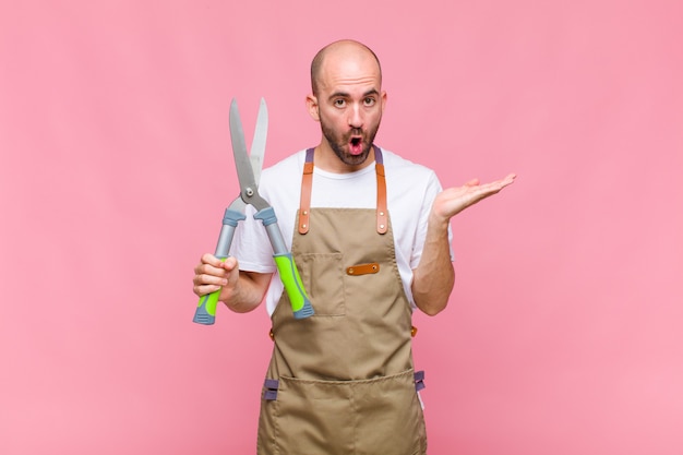 Young bald man looking surprised and shocked, with jaw dropped holding an object with an open hand on the side