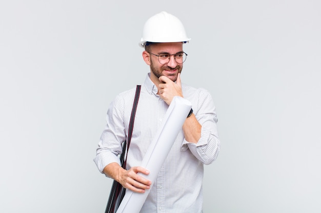 Young bald man smiling with a happy, confident expression with hand on chin, wondering and looking to the side