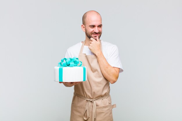 Young bald man smiling with a happy, confident expression with hand on chin, wondering and looking to the side