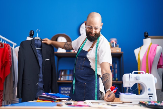 Young bald man tailor smiling confident holding jacket writing on notebook at clothing factory