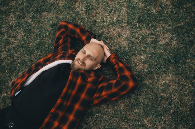 Photo young bald man with a beard lying on the grass in the park and smiling looking at the camera