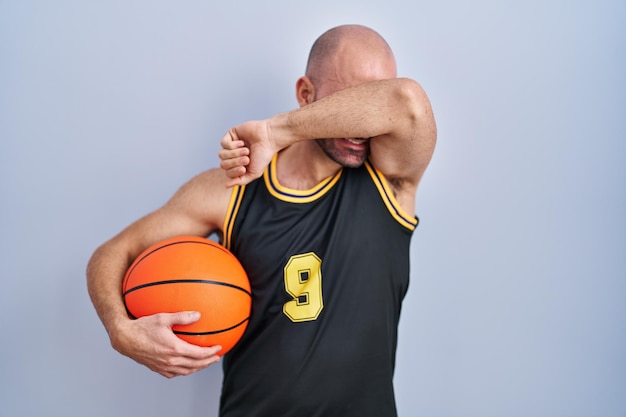 Фото Молодой лысый мужчина с бородой в баскетбольной форме держит мяч, весело улыбаясь, играя в прятки с руками, показывающими лицо. удивлен и вышел