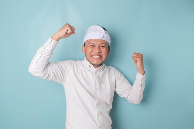 A young Balinese man with a happy successful expression wearing udeng or traditional headband and white shirt isolated by blue background