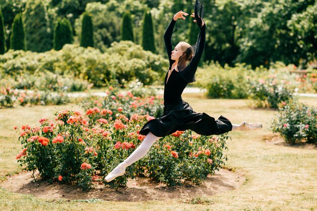 Young ballerina in black dress jumping among rose bushes in park