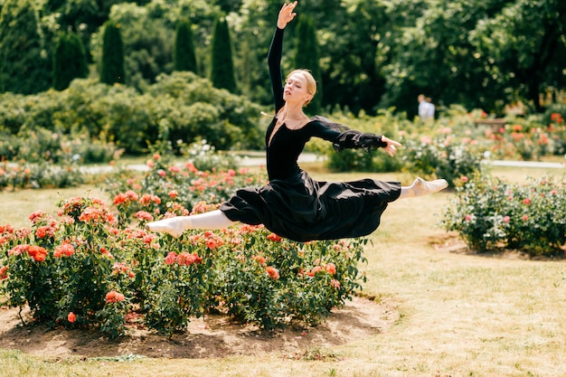 Young ballerina in black dress jumping among rose bushes in park