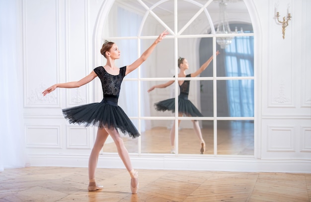 Юная балерина в черной пачке стоит в изящной позе на пуантах в большом светлом зале перед зеркалом.