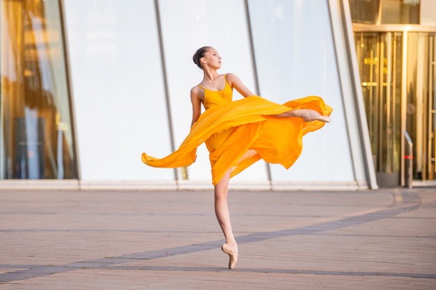 A young ballerina in a long flying yellow dress is dancing against the backdrop of a cityscape