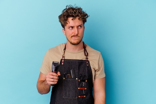 Young barber caucasian man isolated on blue wall confused, feels doubtful and unsure.