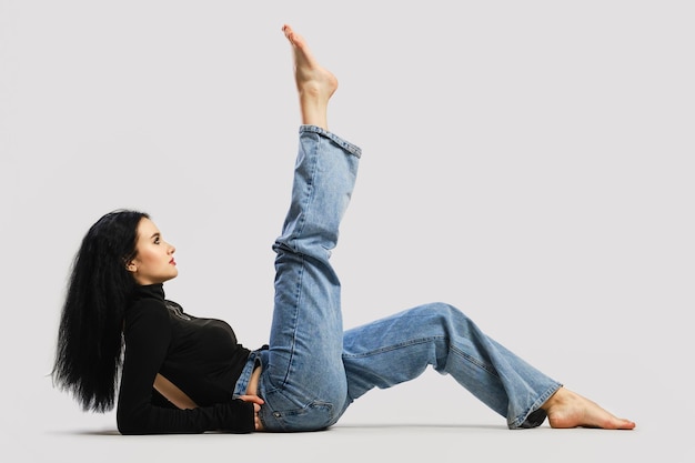 Young barefoot woman lying on the floor and raises one leg up in the studio over grey background