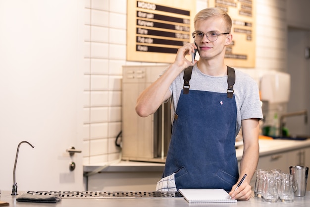 Young barista in apron and t-shirt taking orders of clients on the phone while standing by workplace and making notes in notepad