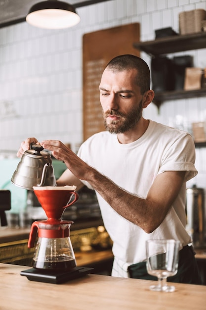 젊은 바리스타는 바 카운터에 서서 카페에서 일하는 동안 커피를 붓고 신중하게 준비합니다.