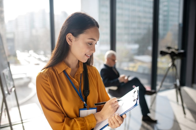 Young beaming pretty asian reporter looking contented while making notes