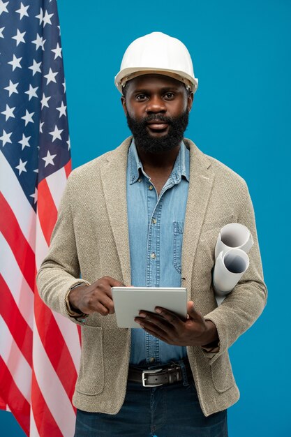 Young bearded African-american engineer using tablet and holding rolled blueprints while standing against stars-and-stripes flag