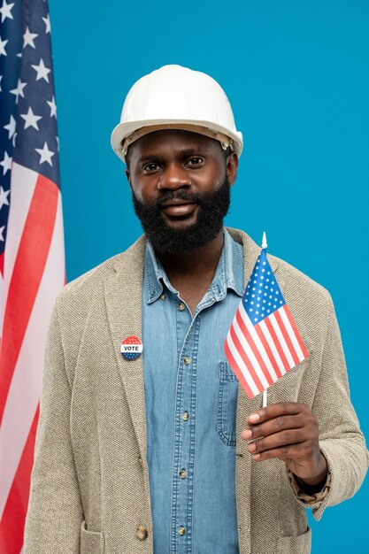 Young bearded African builder or engineer in helmet and formalwear holding small us flag while standing against stars-and-stripes